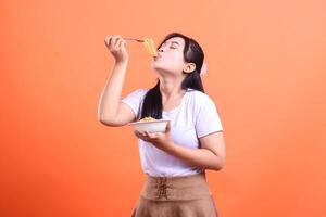A young woman is happily eating noodles from a bowl with a fork, looking satisfied with closed eyes, isolated on orange background photo