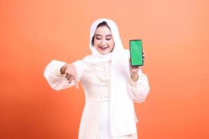 A Muslim woman in hijab smiles while presenting a smartphone with a green screen, pointing down with her other hand to attract attention, isolated on a orange background. photo