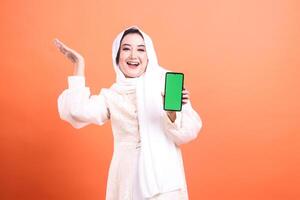 Cheerful Muslim woman in hijab presenting a green screen mobile phone with a happy expression. isolated on a orange background photo
