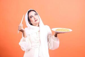 A thoughtful Asian Muslim woman wearing a hijab holding a plate of noodles and chopsticks. isolated on a orange background photo