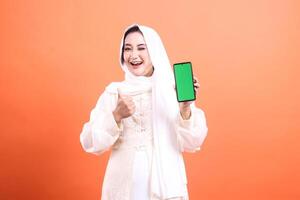 A cheerful Muslim woman in hijab holds up a smartphone with a green screen, giving a thumbs up, and smiling. isolated on a orange background photo