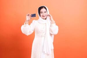 Smiling Muslim Woman Holding Credit Card on Orange Background, showcasing secure and convenient payment methods, isolated on a orange background. photo