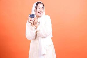 Delighted Muslim Woman Holding Credit Card A bright and cheerful portrait of financial empowerment and modern lifestyle, isolated on a orange background. photo