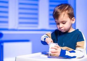 Child enjoys yogurt in bright room, a young child tastes yogurt from a small bowl, sitting at a white table in a well-lit room with blue tones. photo
