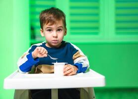 Child enjoying a snack at a table. A young boy sits at a table spooning yogurt, with green walls surrounding him in a playful atmosphere. photo