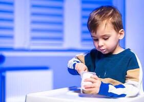 Child enjoys yogurt in bright room. A young child tastes yogurt from a small bowl, sitting at a white table in a well-lit room with blue tones. photo