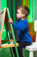 Child happily drawing creatively. A child in a blue shirt sits at an easel, focused on drawing with colorful materials in a vibrant indoor setting. photo