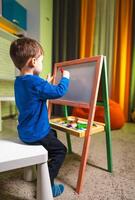 Kid drawing at easel inside. A child focused on creating art at a mini easel in a vibrant indoor space during the day. photo