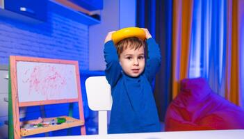 Child plays with a yellow toy on head. A young boy wearing a blue top holds a yellow toy above his head in a brightly colored room. photo