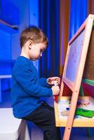 Child drawing creatively. A child is focused on drawing with markers at an easel in a brightly lit room, showcasing creativity and concentration. photo