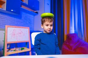 Child balances green item indoors. A young child sits at a table with a green object balanced on their head in a colorful indoor setting. photo