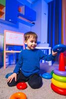 Kid plays with stacking toys. A cheerful young boy is stacking colorful rings on a cone in a well-lit playroom filled with toys. photo