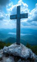 A wooden cross is on a rock in the mountains. The sky is blue and cloudy. The mountains are in the background and there is a lake in the distance photo