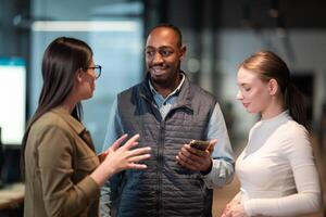 African American male coach or speaker sharing business ideas and collaborating with a diverse team in a modern office, presenting a project on a smartphone during a brainstorming session in a startup photo