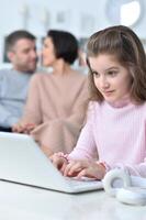 Cheerful little girl using a laptop at home with her parents in the background. photo