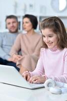 Cheerful little girl using a laptop at home with her parents in the background. photo