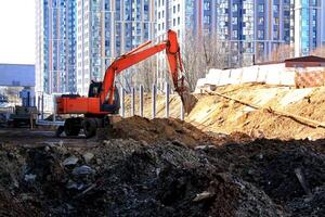 Orange excavator digging at a construction site, with a large building under construction in the background. Construction equipment, urban development, and infrastructure project photo
