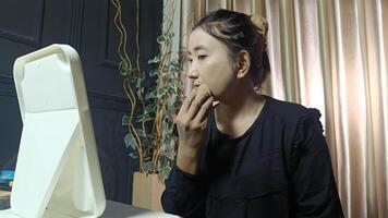Close-up of an Asian woman applying foundation to her face using a makeup sponge. She is looking in a mirror in her home. video