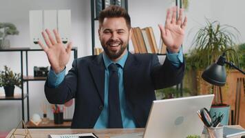 Businessman working on laptop smiling friendly at camera and waving hands gesturing hello at office video