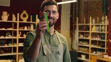 Happy Caucasian man male businessman small business owner potter looking camera hold mobile phone empty screen cellphone chroma key mock up ads showing thumb up approval gesture recommend in workshop video