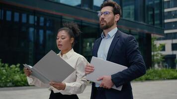 Confident Caucasian man businessman male guy with African American female businesswoman girl business partners talking walking in city downtown hold clipboard with documentation discussing meeting video