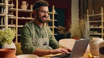 Happy joyful smiling Caucasian man small business owner craftsman businessman potter crock male working laptop typing desktop browsing computer online shopping pottery selling inside workshop studio video