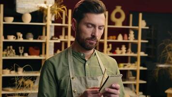 Happy cheerful Caucasian man male small business owner potter entrepreneur businessman writing notes notebook write thinking having idea eureka insight inspiration creativity indoors pottery workshop video