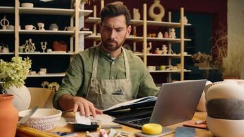 Serious frowned Caucasian man male small business owner businessman potter craftsman counting expense payment calculator taxes working laptop look notebook writing accounting indoors pottery workshop video