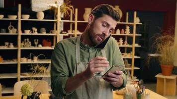 Happy Caucasian man male guy small business owner businessman potter craftsman multitasking writing notes write plan talking mobile phone call chat client customer conversation inside pottery workshop video