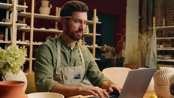 Happy smiling Caucasian man guy small business owner businessman successful entrepreneur crock potter artist male working computer typing laptop internet website looking camera inside pottery workshop video
