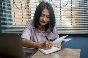 Woman sitting on chair writing note while using laptop at table study online course development program, working at home reskill upskill work concepts photo