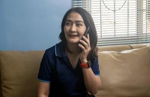 Asian woman sitting on sofa using mobile smart phone talking with friend, conversation chatting relaxing lifestyle concepts photo