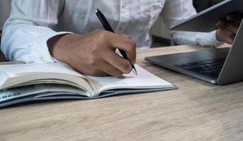 Men hand using pen writing on paper notebook homework on table and holding tablet search information education,Student online learning concepts photo