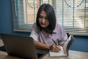 Woman sitting on chair writing note while using laptop at table study online course development program, investment reskill upskill work concepts photo