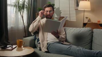 Caucasian man reading book alone at home male homeowner reader smart guy businessman in eyeglasses read literature on couch sofa paper pages textbook hobby leisure rest in apartment studying knowledge video
