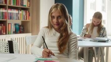 Cute schoolgirl kid girl student Caucasian teen child gen z pupil learner at classroom writing task exam learn lesson exercise smiling teenager looking at camera education knowledge studying in school video