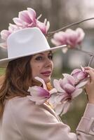 Woman magnolia flowers, surrounded by blossoming trees., hair down, white hat, wearing a light coat. Captured during spring, showcasing natural beauty and seasonal change. photo