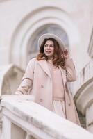 woman in elegant coat and hat against an intricate architectural backdrop, harmoniously blending modern fashion with historical allure. The soft daylight adds to its timeless appeal. photo