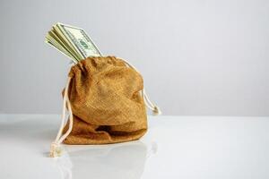 A small, brown burlap bag sits on a white surface. The bags drawstring is pulled tight, and a bundle of US dollar bills is peeking out from the top photo
