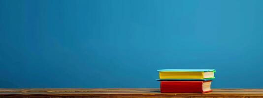 A stack of vibrant books in various colors is placed on a wooden table, creating an inviting atmosphere in a warm and cozy space with a blue backdrop photo