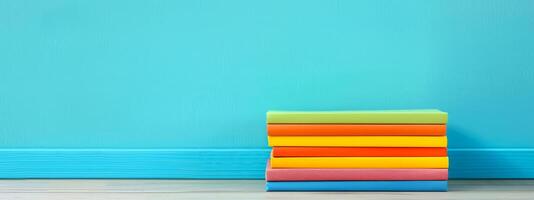 A stack of seven brightly colored books arranged neatly on a light wooden table, creating a lively contrast against a deep blue wall, inviting reading photo