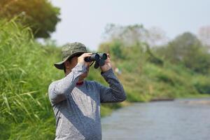 Asian man uses binoculars to observe local birds along a river. photo