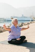 Serious aged woman in with dreadlocks hairstyle standing on beach against sea while practicing pose or asana during sunny day photo