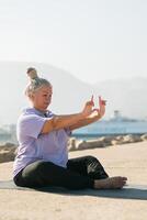 Serious aged woman in with dreadlocks hairstyle standing on beach against sea while practicing pose or asana during sunny day photo