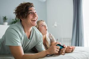 Young couple in love playing games while lying on the bed in the bedroom, having fun and enjoying each other on their weekends. Technologies and gaming concept photo