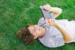 Joyful man smiling and having fun while playing games with portable console and lying on the grass outdoors. Technology and game addiction concept photo