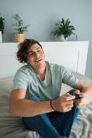 Handsome excited gamer sitting on a bed and playing games on a console. Wireless controller and online game technologies concept photo