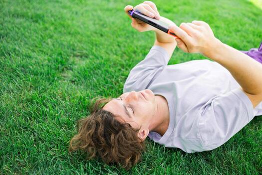 Joyful man smiling and having fun while playing games with portable console and lying on the grass outdoors. Technology and game addiction concept photo
