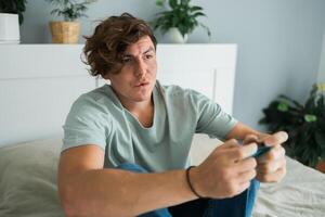 Handsome excited gamer sitting on a bed and playing games on a console. Wireless controller and online game technologies concept photo
