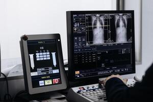 In Control Room Doctor and Radiologist Discuss Diagnosis while Watching Procedure and Monitors Showing Lungs Scans Results, In the Background Patient Undergoes MRI or CT Scan Procedure photo